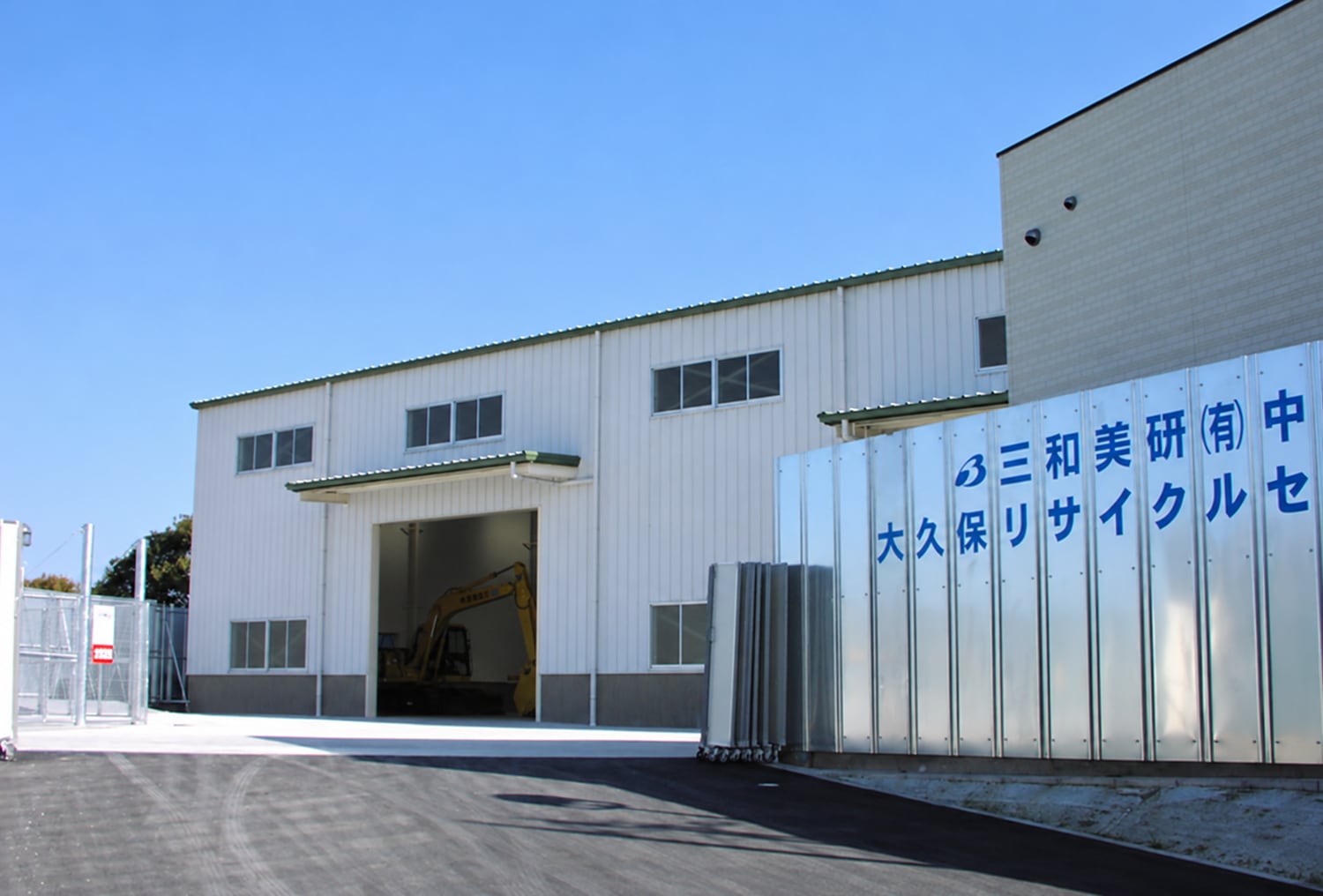 大久保リサイクルセンター。青空の下に白い処理棟があり、開いた入口の奥に黄色い重機が見える。右に社名とセンター名の看板。