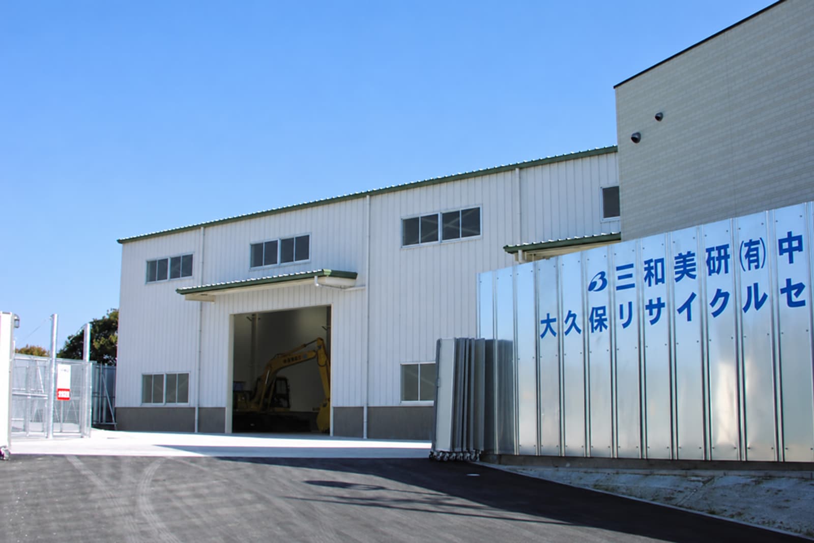 大久保リサイクルセンター。青空の下に白い処理棟があり、入口の奥に黄色い重機が見える。右に社名とセンター名の看板。
