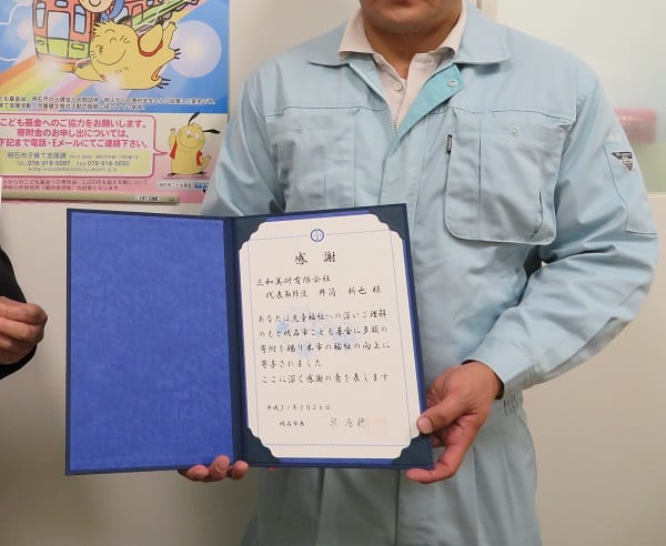 明石市長から三和美研有限会社に贈られた、明石市こども基金への寄付に対する感謝状を手に持つ様子。後ろの壁に、こども基金への協力を呼びかけるポスター。